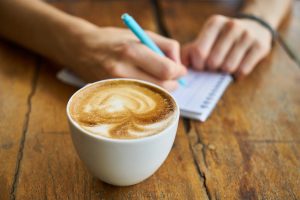 Eine Tasse Kaffee mit geschäumter Milch, im Hintergrund ist eine Person und notiert etwas auf einem Blog.